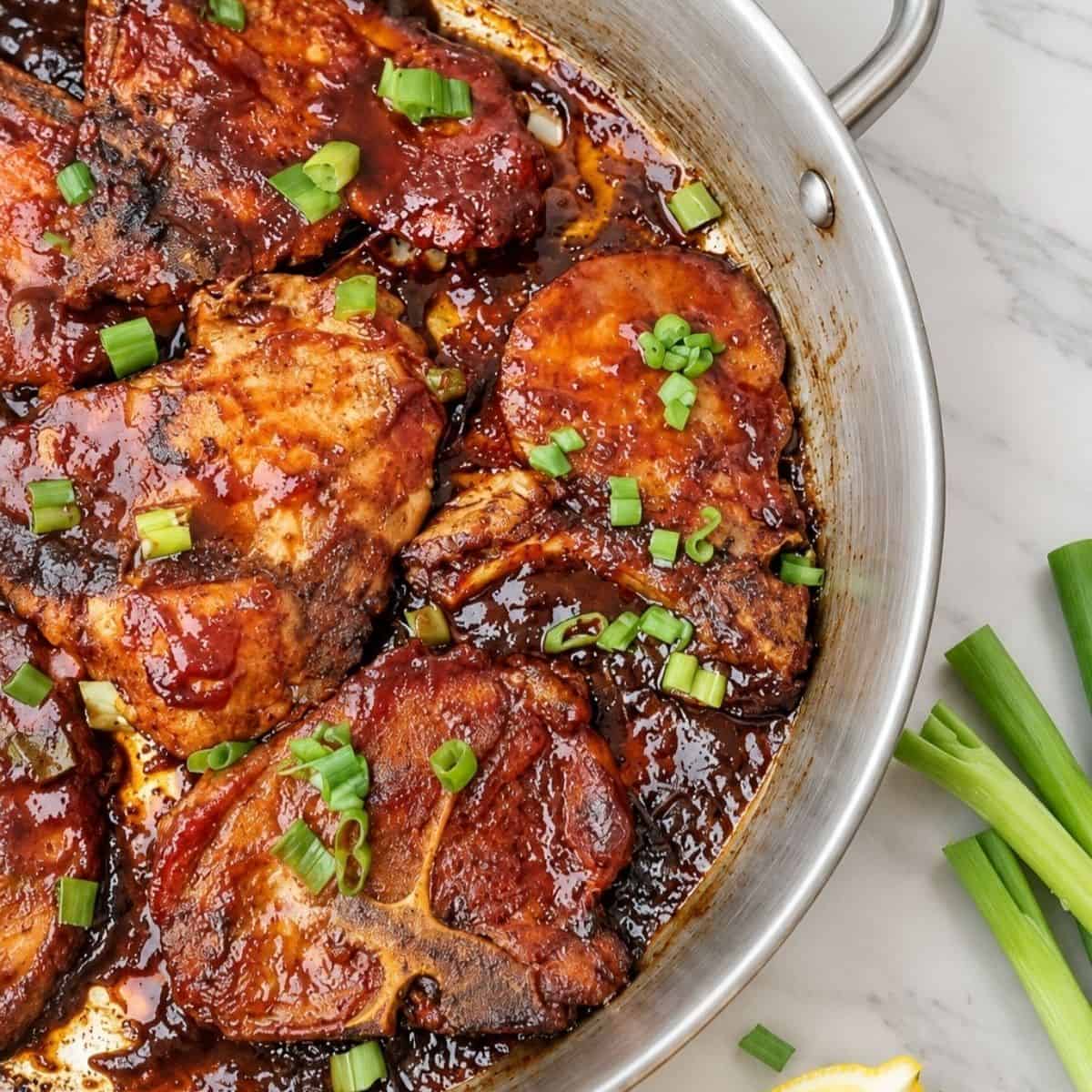 Pork chops coated in barbecue sauce with chopped green onions in a stainless steel skillet.