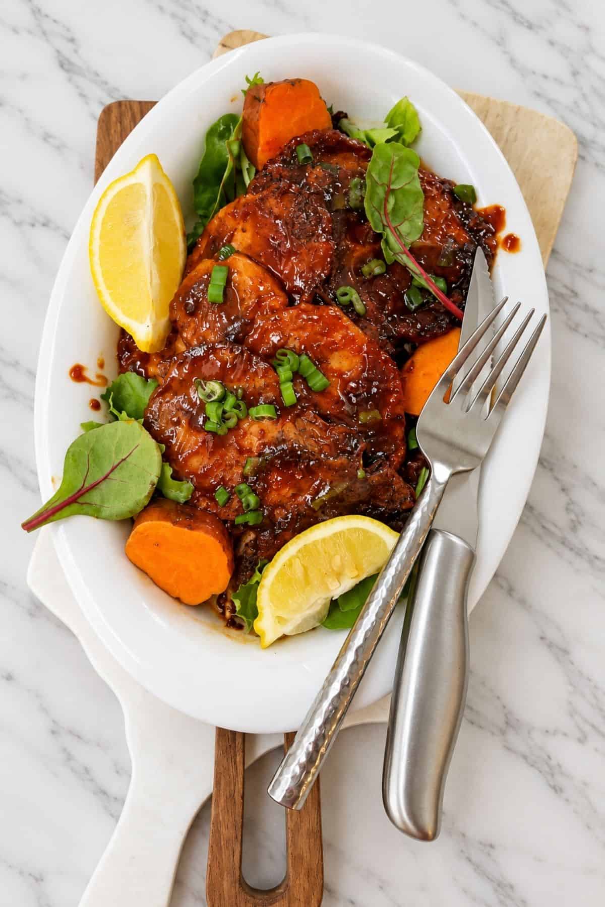 Pan-fried BBQ pork chops on a white serving platter with carrot pieces, leafy greens, lemon wedges, and a fork and knife.
