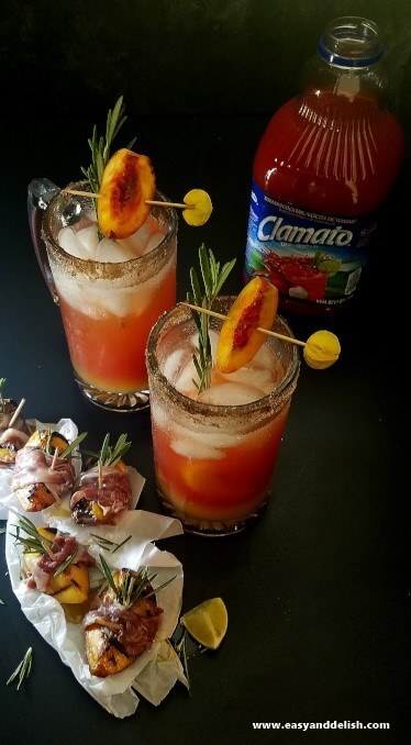 Two beer glass mugs filled with peach rosemary michelada between a bottle of juice and skewers of peach wrapped with prosciutto. 