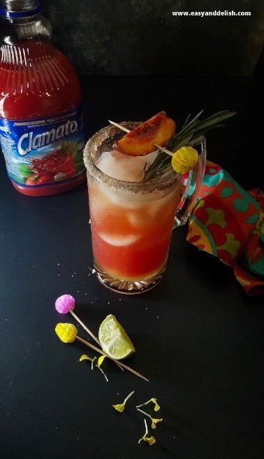 A beer glass mug of peach rosemary michelada between a bottle of Clamato tomato juice and a wedge of lime. 