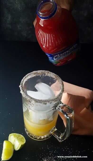 A bottle of Clamato tomato juice being poured into a beer glass mug filled with ice and peach juice. 