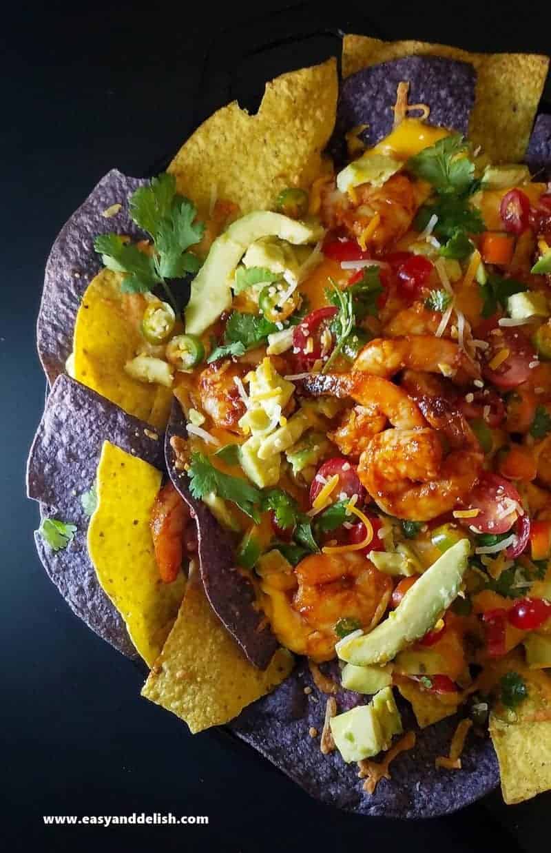 Overhead view of colorful tortilla chips layered on a skillet topped with nacho sauce, shrimp, avocado, and salsa. 
