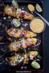 Four ears of nacho grilled corn on the cob and a bowl of sauce on a baking sheet