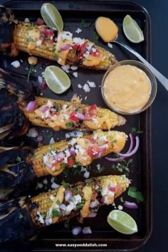 Four ears of nacho grilled corn on the cob and a bowl of sauce on a baking sheet