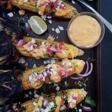 Four ears of nacho grilled corn on the cob and a bowl of sauce on a baking sheet