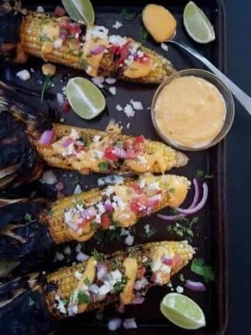 Four ears of nacho grilled corn on the cob and a bowl of sauce on a baking sheet