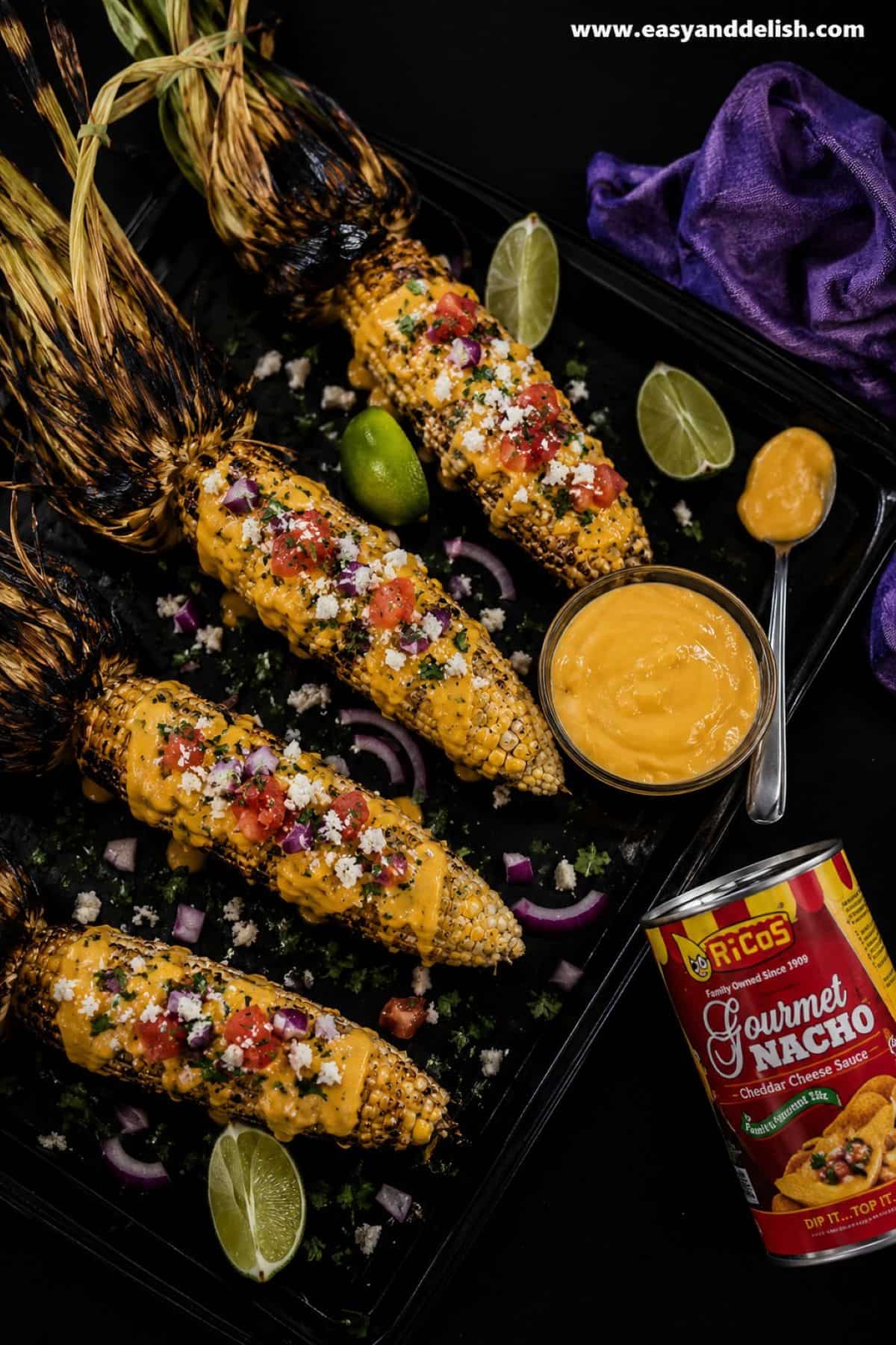 A quick and easy Nacho Grilled Corn on the Cob topped with nacho sauce, pico de gallo, and queso fresco or feta. The perfect side to serve at bbq's and summer backyard parties. Grilled nacho corn on the cob topped with creamy cheddar cheese sauce, crumbled cotija cheese, diced tomatoes, red onion, fresh cilantro, and served with a lime wedge on a dark baking tray.