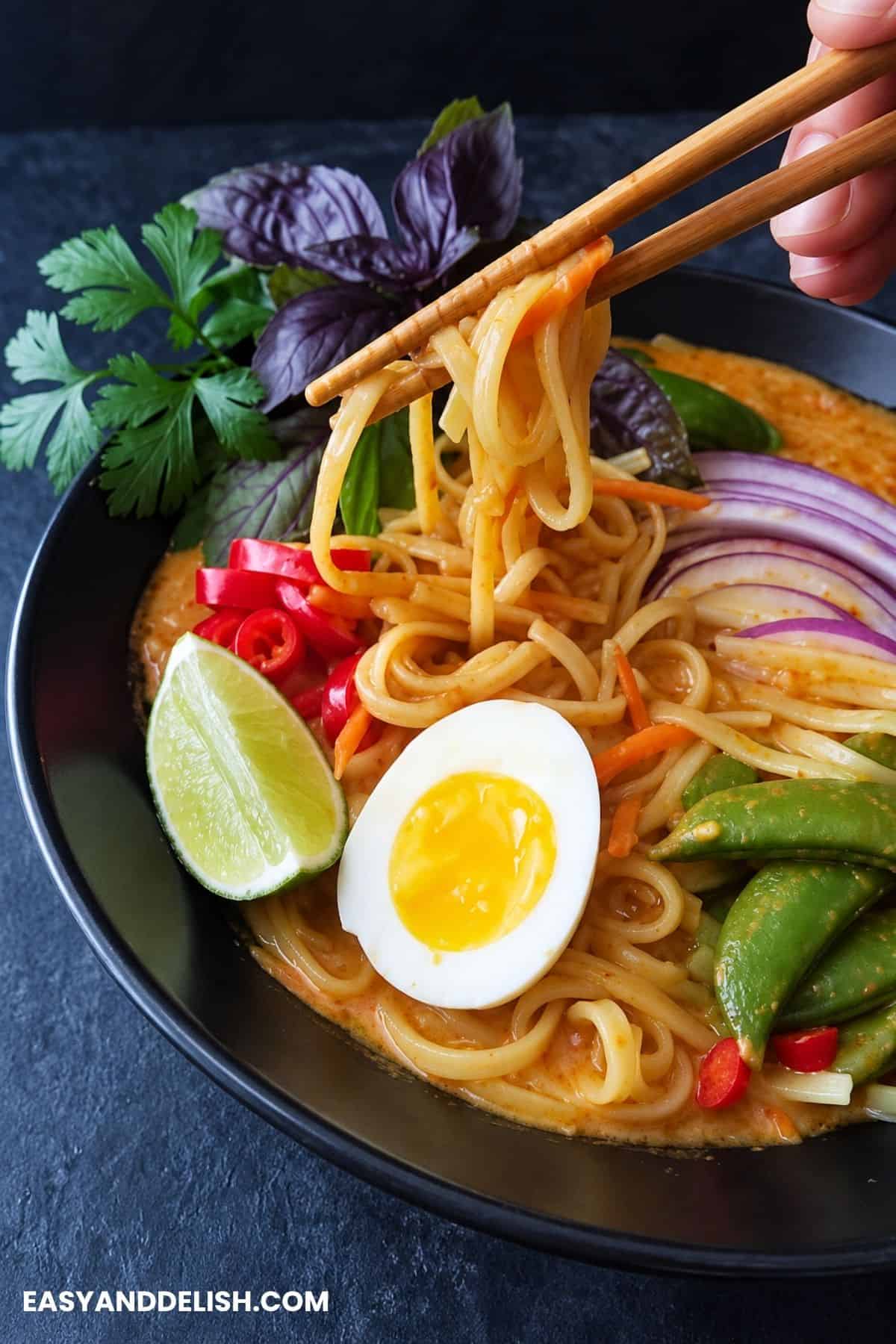 Rice noodles lifted with chopsticks from coconut curry broth with snap peas, peppers, onion, herbs, lime, and egg.