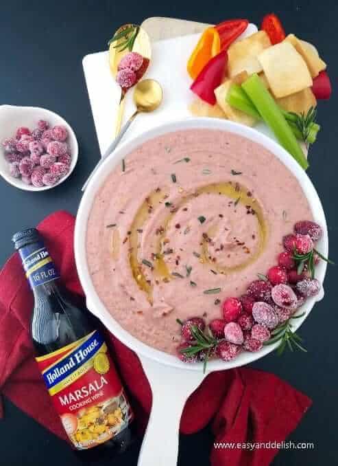 A bowl of cranberry dip with some crudite vegetables on the side and  garnishes. 