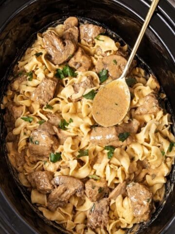Beef and egg noodles in creamy mushroom sauce inside a slow cooker with a serving spoon.