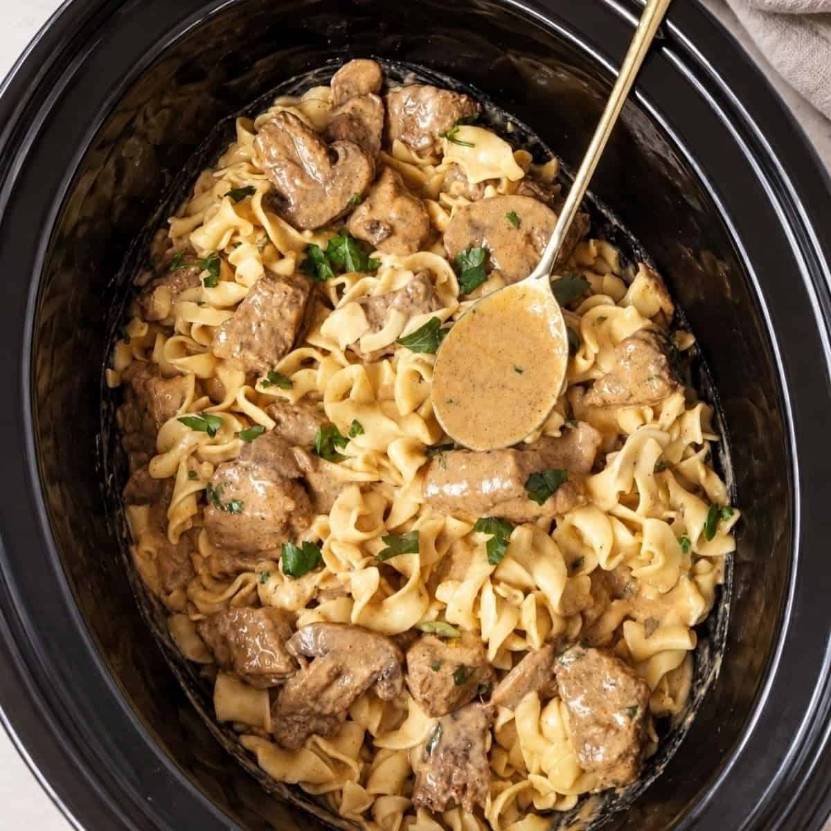 Beef and egg noodles in creamy mushroom sauce inside a slow cooker with a serving spoon.