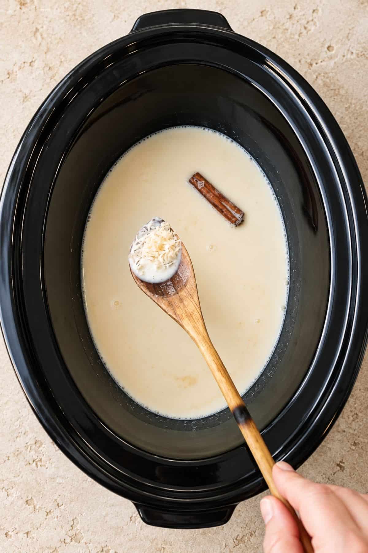 Arborio rice on a wooden spoon above milk and a cinnamon stick in a slow cooker.
