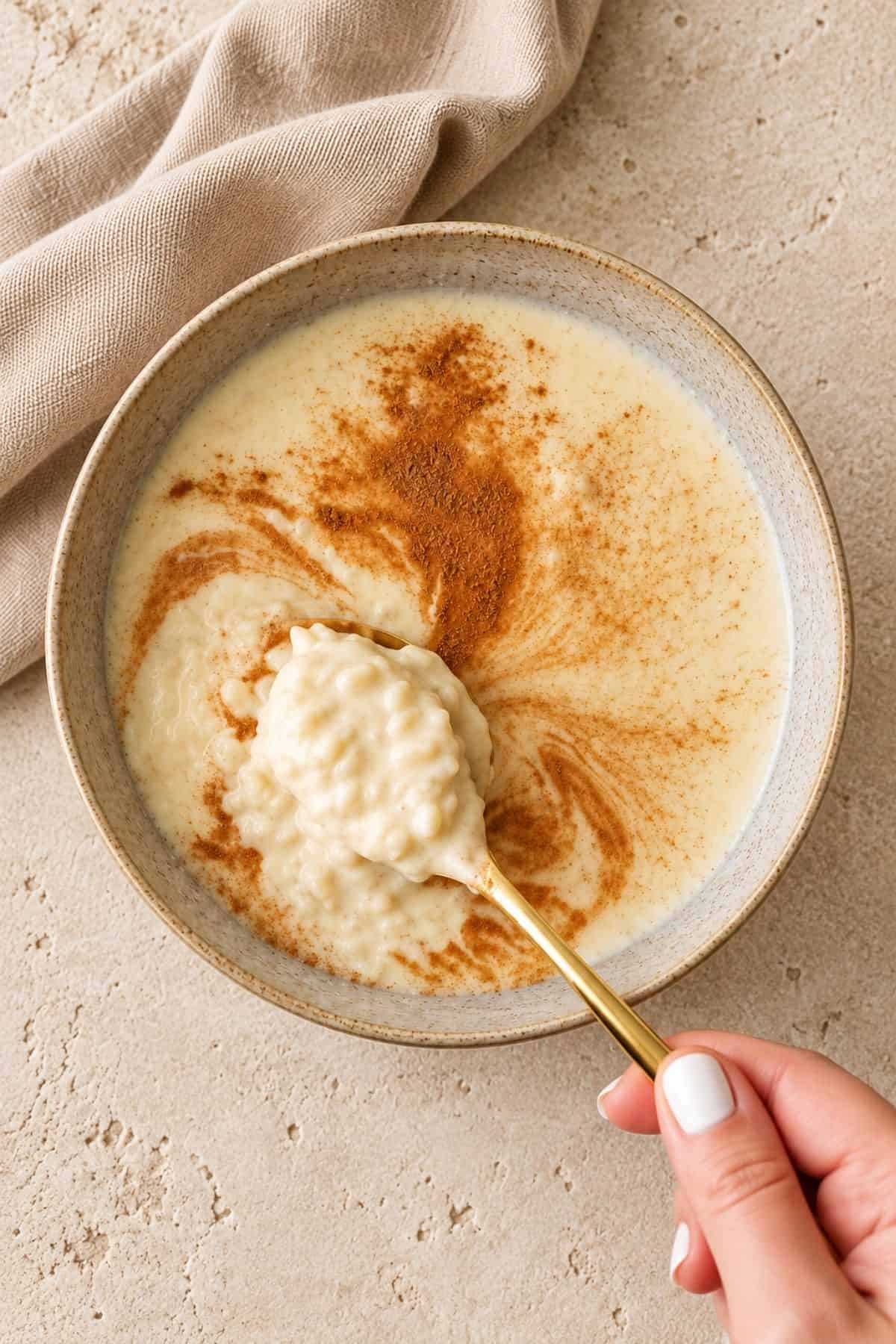 A close-up of a spoonful of Slow Cooker Rice Pudding above a gray bowl with cinnamon swirled into the creamy mixture.