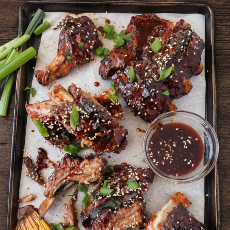 Char siu ribs topped with sesame seeds and green onions on a parchment-lined baking sheet beside a bowl of sauce.