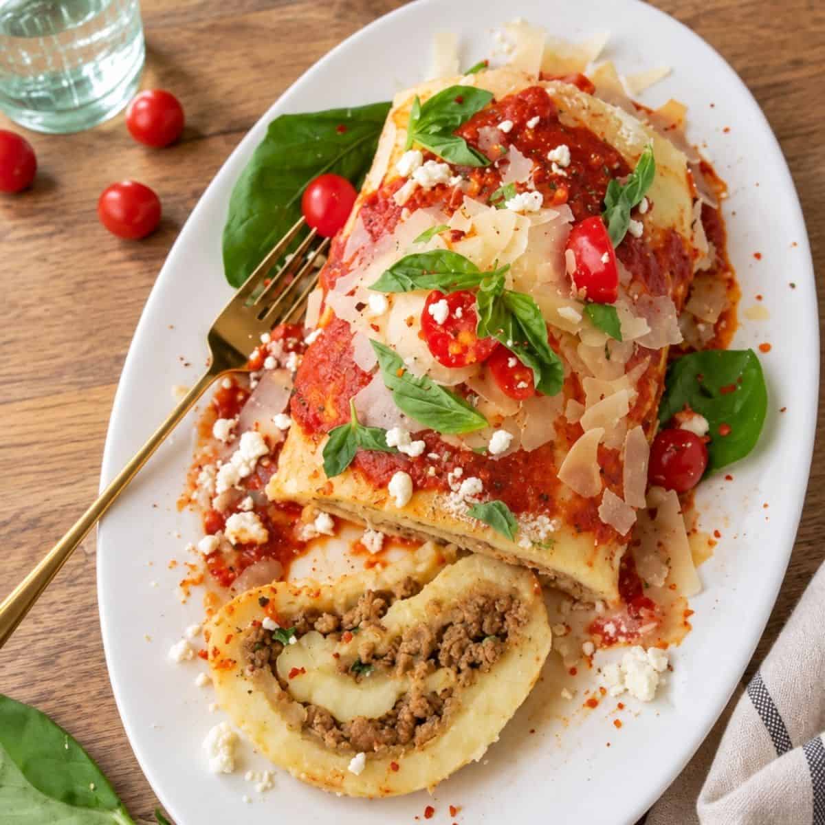 Potato roulade on a white platter with tomato sauce, Parmesan, basil, and a slice filled with beef and cheese.