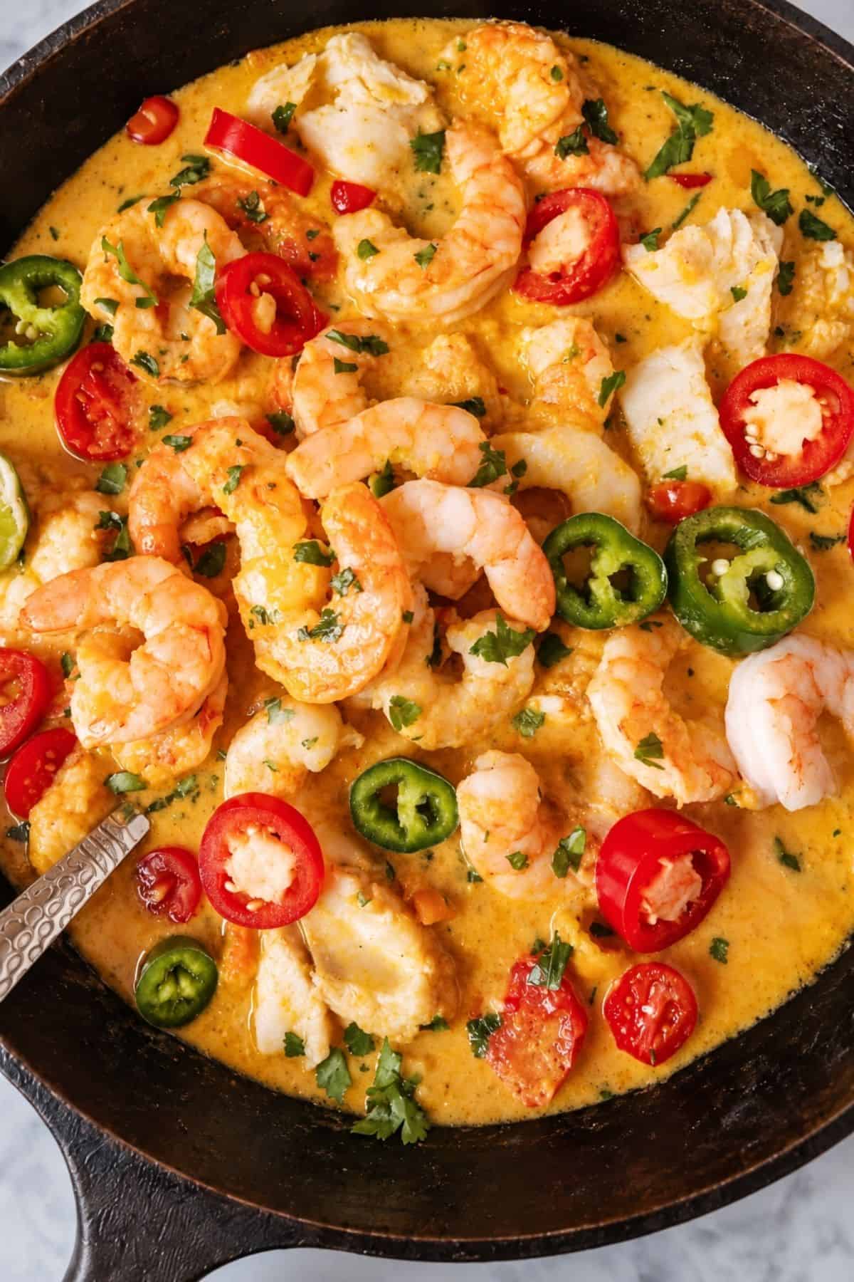 Close-up of fish and shrimp in a creamy orange coconut sauce with sliced red and green chiles and chopped herbs.