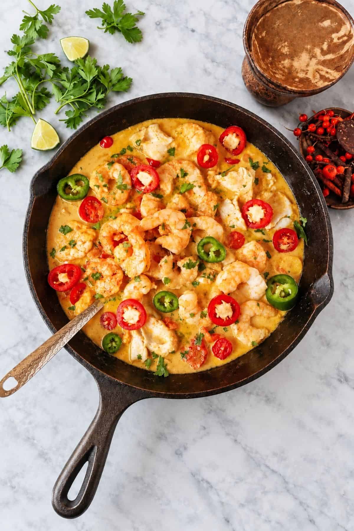 Screenshot of a design editor showing an overhead view of moqueca with shrimp, fish, sliced chiles, and an ornate spoon in a skillet.