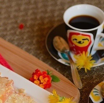 Table scene with a cup of coffee that has a smiling red and yellow sloth painted on it