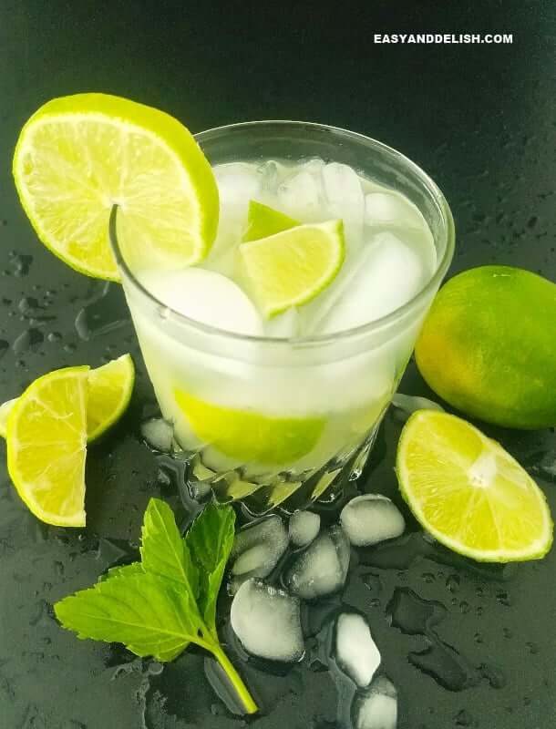 Clear glass filled with caipirinha drink, ice cubes, and wedges of lime, garnished with a slice of lime on the rim of the glass