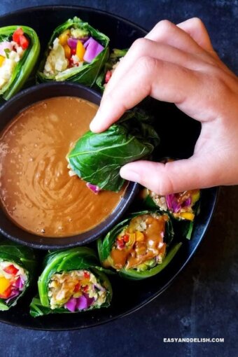 A bowl of collard green wraps with a small bowl of peanut sauce in the middle and one of them being dipped