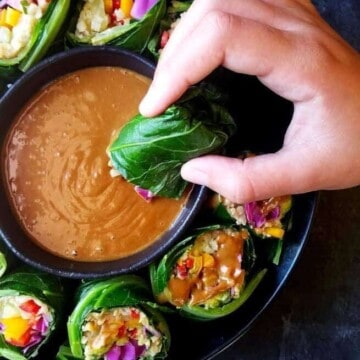 A bowl of collard green wraps with a small bowl of peanut sauce in the middle and one of them being dipped