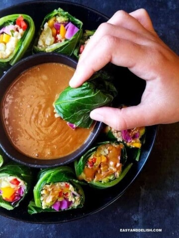 A bowl of collard green wraps with a small bowl of peanut sauce in the middle and one of them being dipped