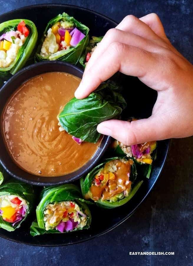 A bowl of collard green wraps with a small bowl of peanut sauce in the middle and one of them being dipped