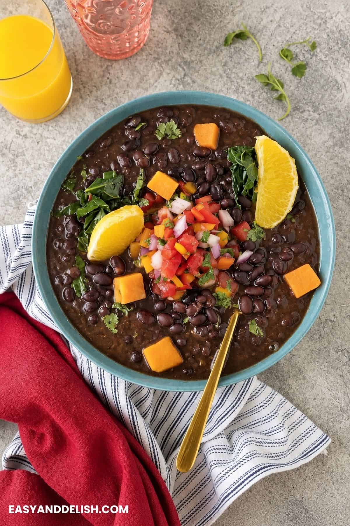 Bowl of vegetarian feijoada topped with sweet potatoes, greens, fresh salsa, cilantro, and orange wedges.
