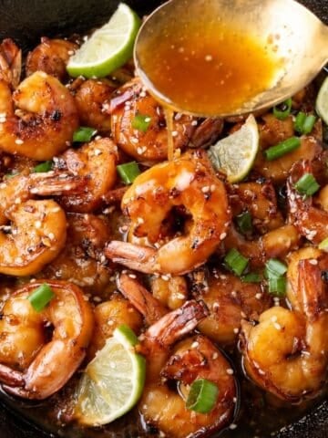A spoon drizzles sauce over seared shrimp garnished with sesame seeds, green onions, and lime wedges in a cast-iron skillet.