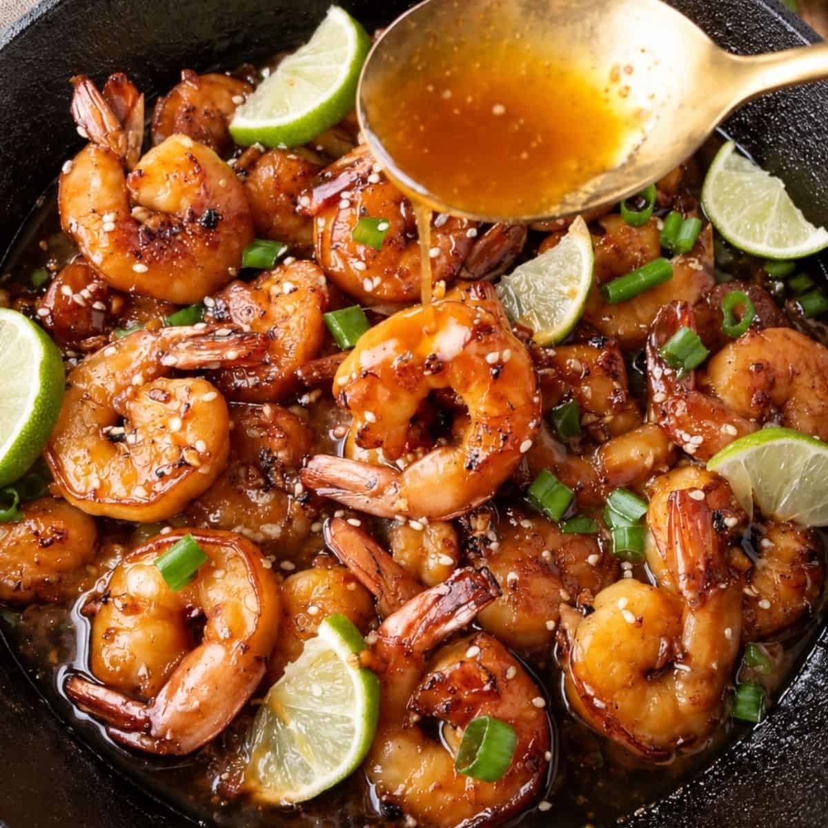 A spoon drizzles sauce over seared shrimp garnished with sesame seeds, green onions, and lime wedges in a cast-iron skillet.