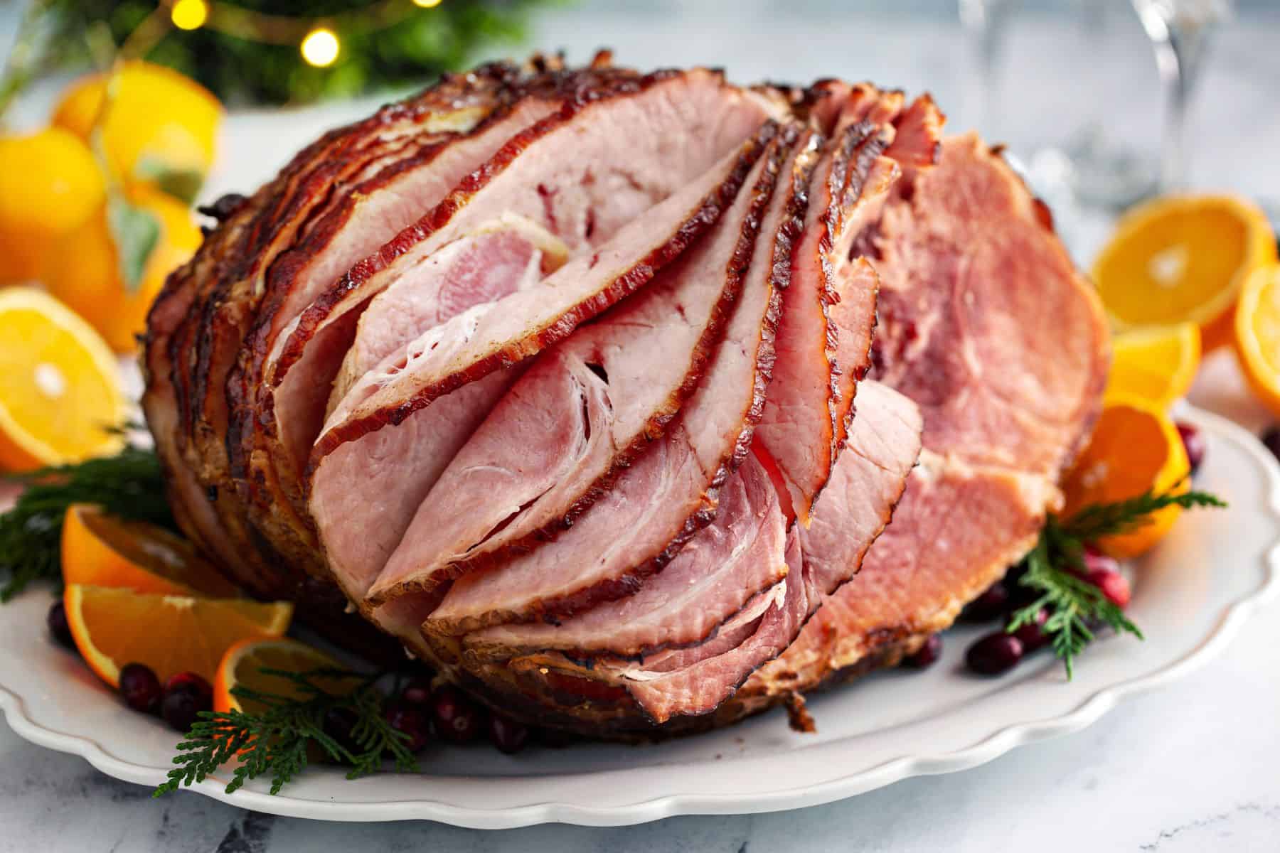 Crockpot spiral ham with orange slices and other garnishes in a platter with holiday decor behind.