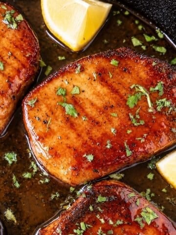 Golden-brown pork chops in a dark skillet with pan sauce, chopped parsley, and lemon wedges.