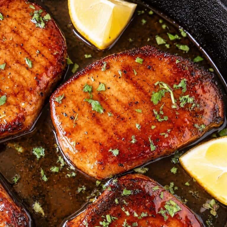 Golden-brown pork chops in a dark skillet with pan sauce, chopped parsley, and lemon wedges.