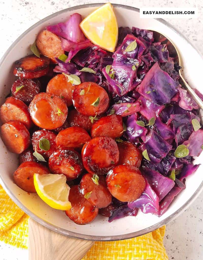 a skillet with smoked sausage and cabbage with lemon wedges and a spoon on the sidei