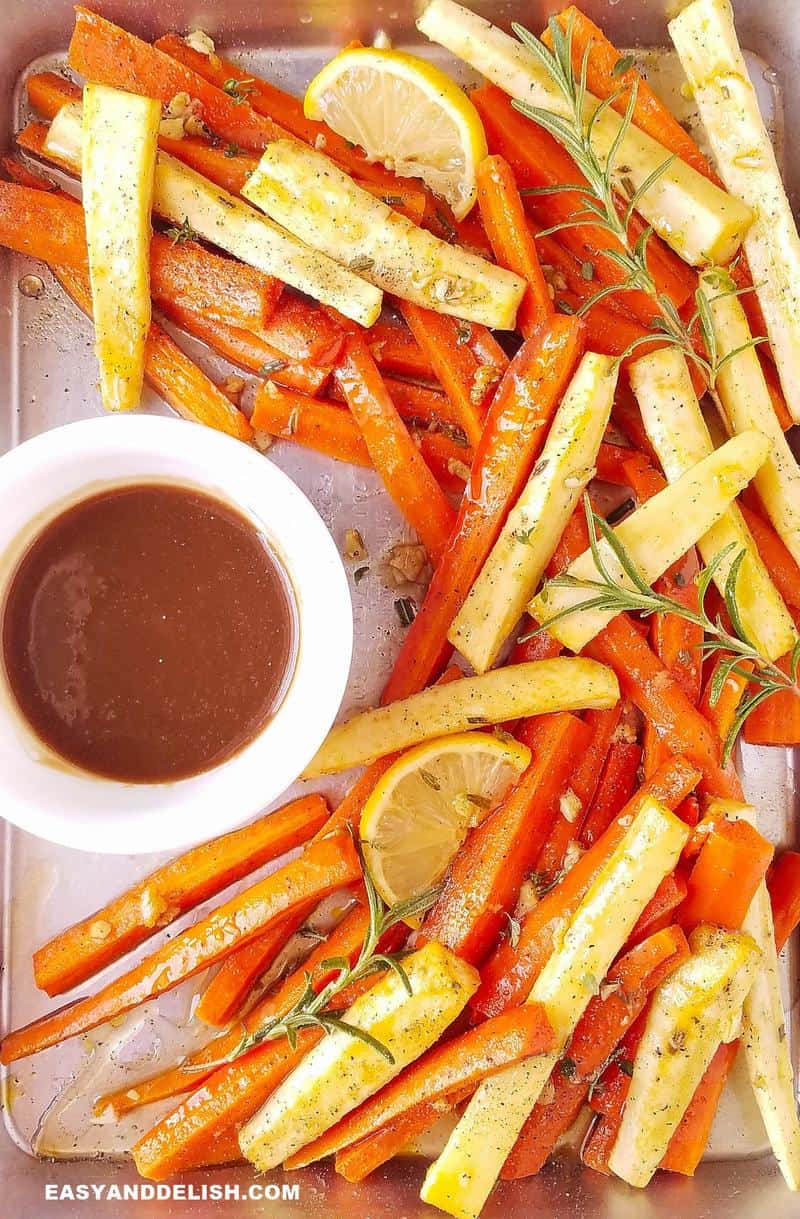 Roasted carrots and parsnips in a baking sheet with a bowl of honey mustard dressing on the side. 