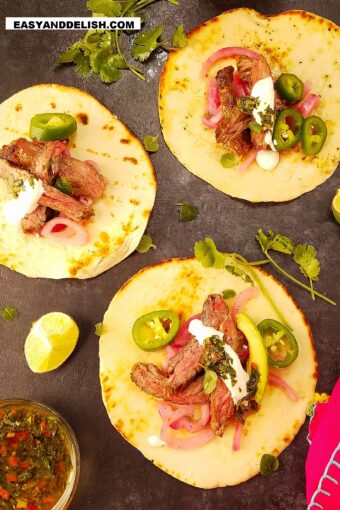 3 carne asada tacos on a surface with garnishes on the side