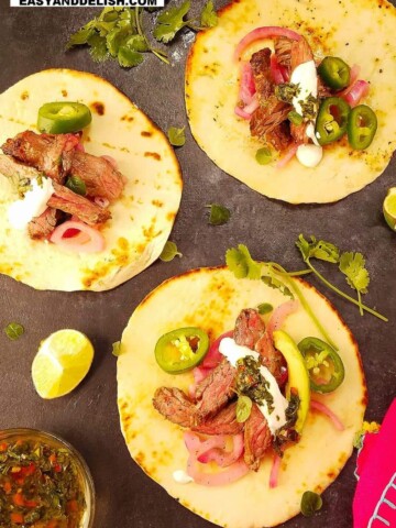 3 carne asada tacos on a surface with garnishes on the side
