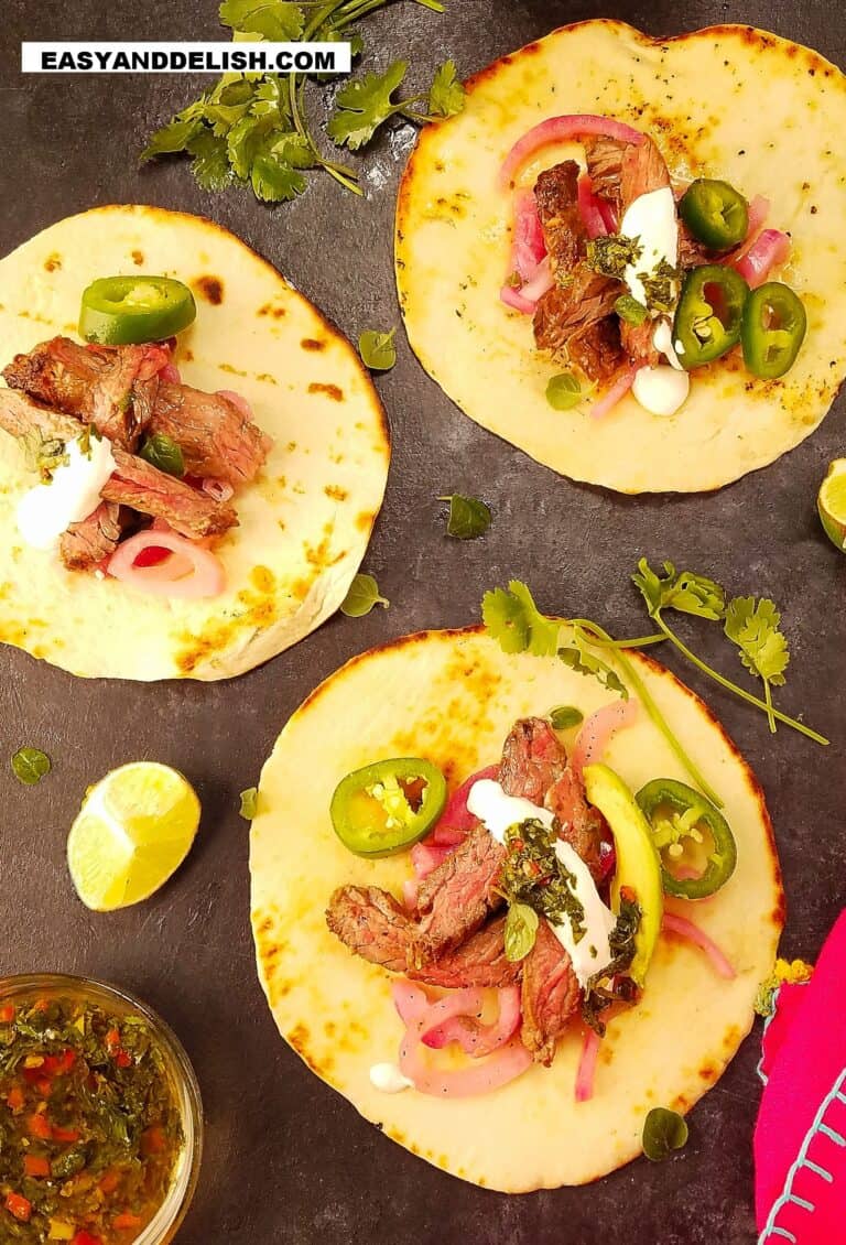 3 carne asada tacos on a surface with garnishes on the side
