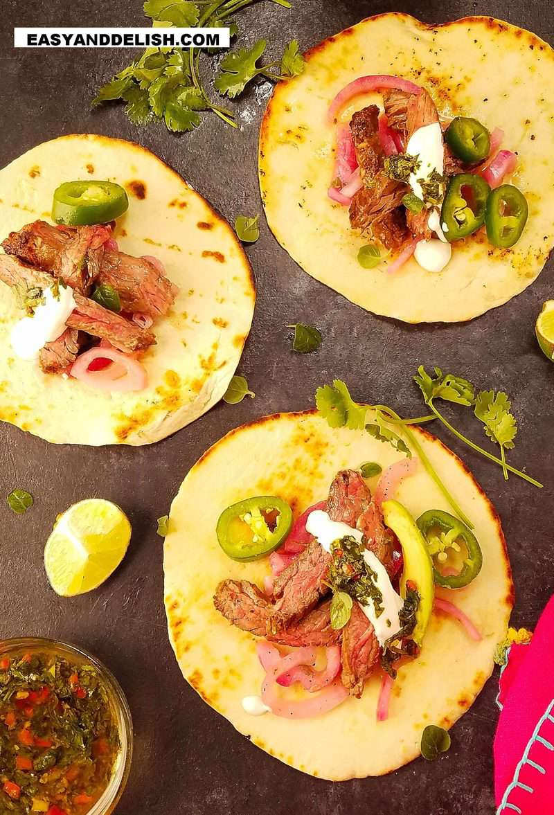 3 carne asada tacos resting on a surface with garnishes on the side