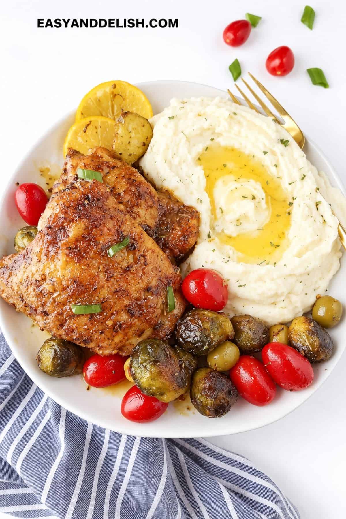 Baked chicken thighs served with mashed potatoes, roasted Brussels sprouts, tomatoes, and olives.