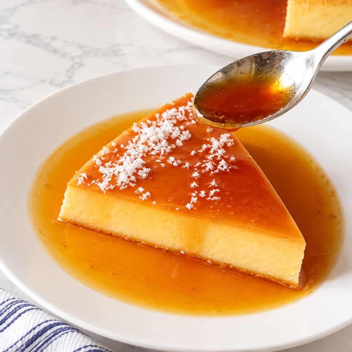 A spoon pours amber caramel sauce over a triangular slice of coconut custard topped with shredded coconut on a plate.