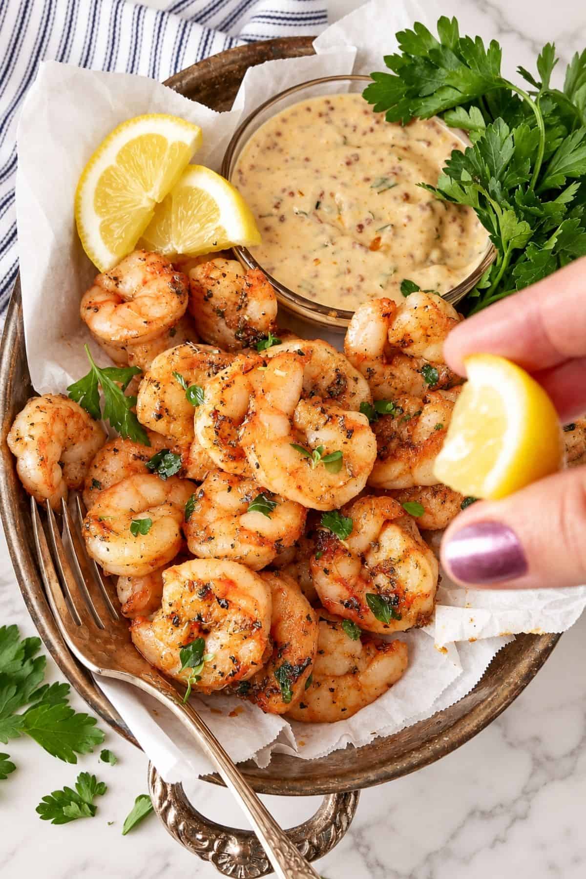 Hand squeezing a lemon wedge over seasoned shrimp beside a bowl of dipping sauce.