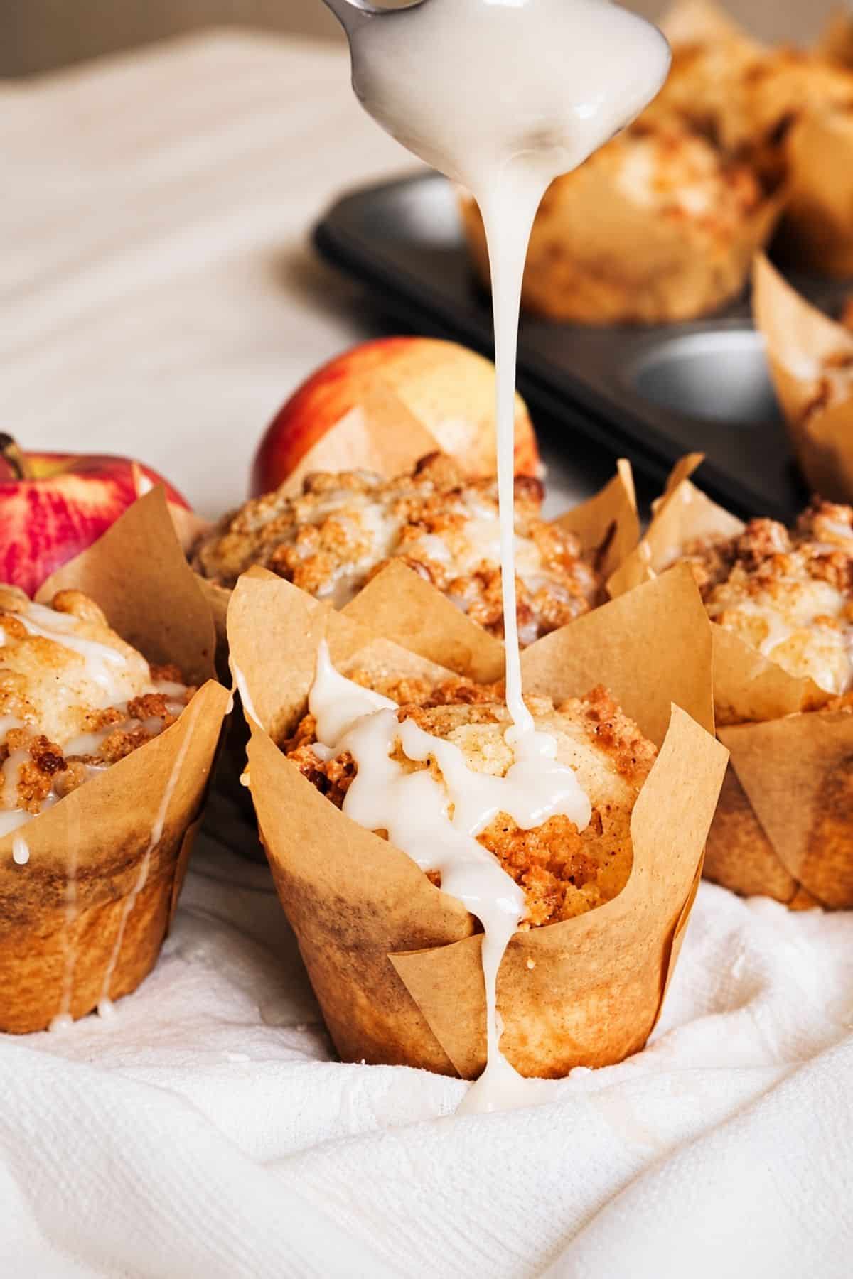 Vanilla glaze spooned over warm apple crumble muffins on a baking pan.