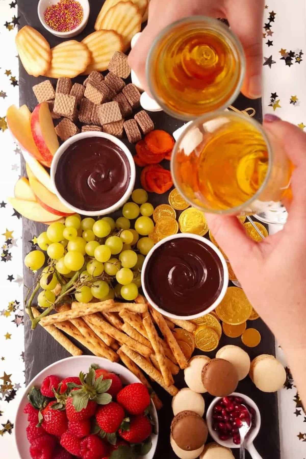 Chocolate fondue board with two glasses of amber drinks, strawberries, grapes, cookies, pretzels, marshmallows, and macarons.