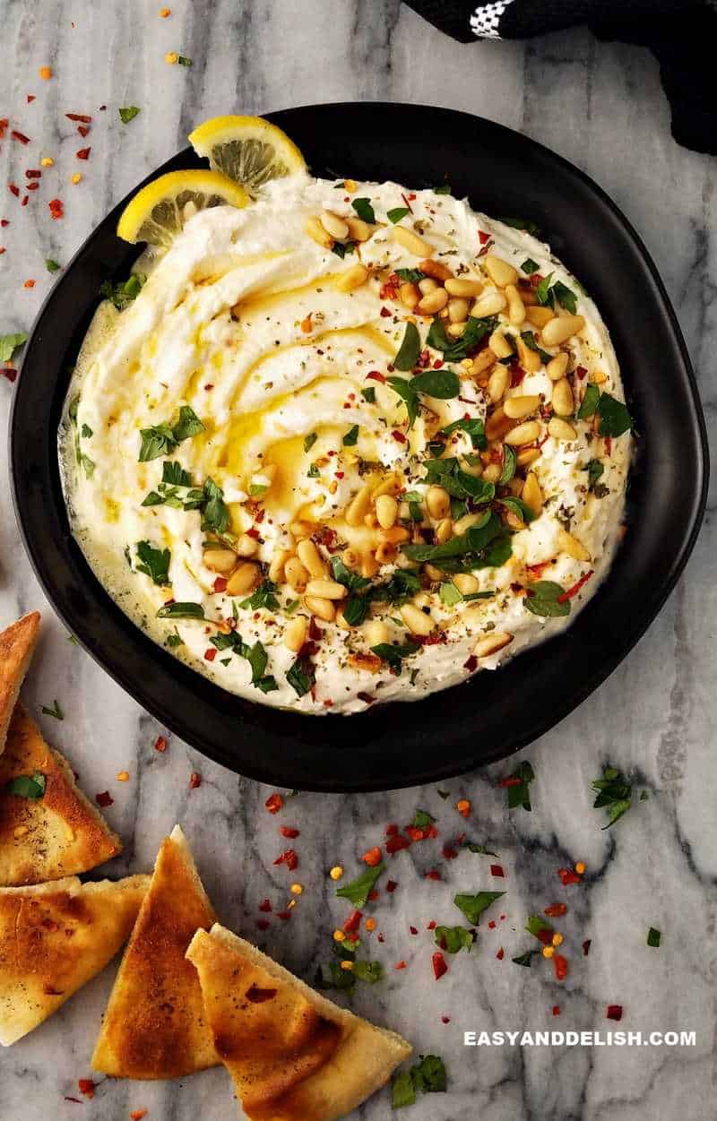 a plate of creamy spreat garnished with olvie oil, honey, pinenuts, herbs and red pepepr flakes with pita chips on the side