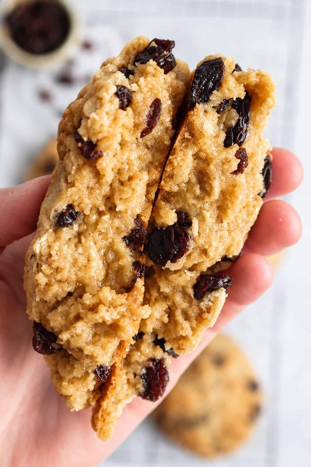 A large oatmeal raisin cookie held in one hand, showing a thick, golden-brown surface with visible raisins and oats.