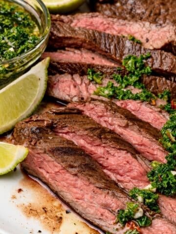 Close-up of pink-centered skirt steak slices topped with chimichurri, with lime wedges and a bowl of sauce alongside.