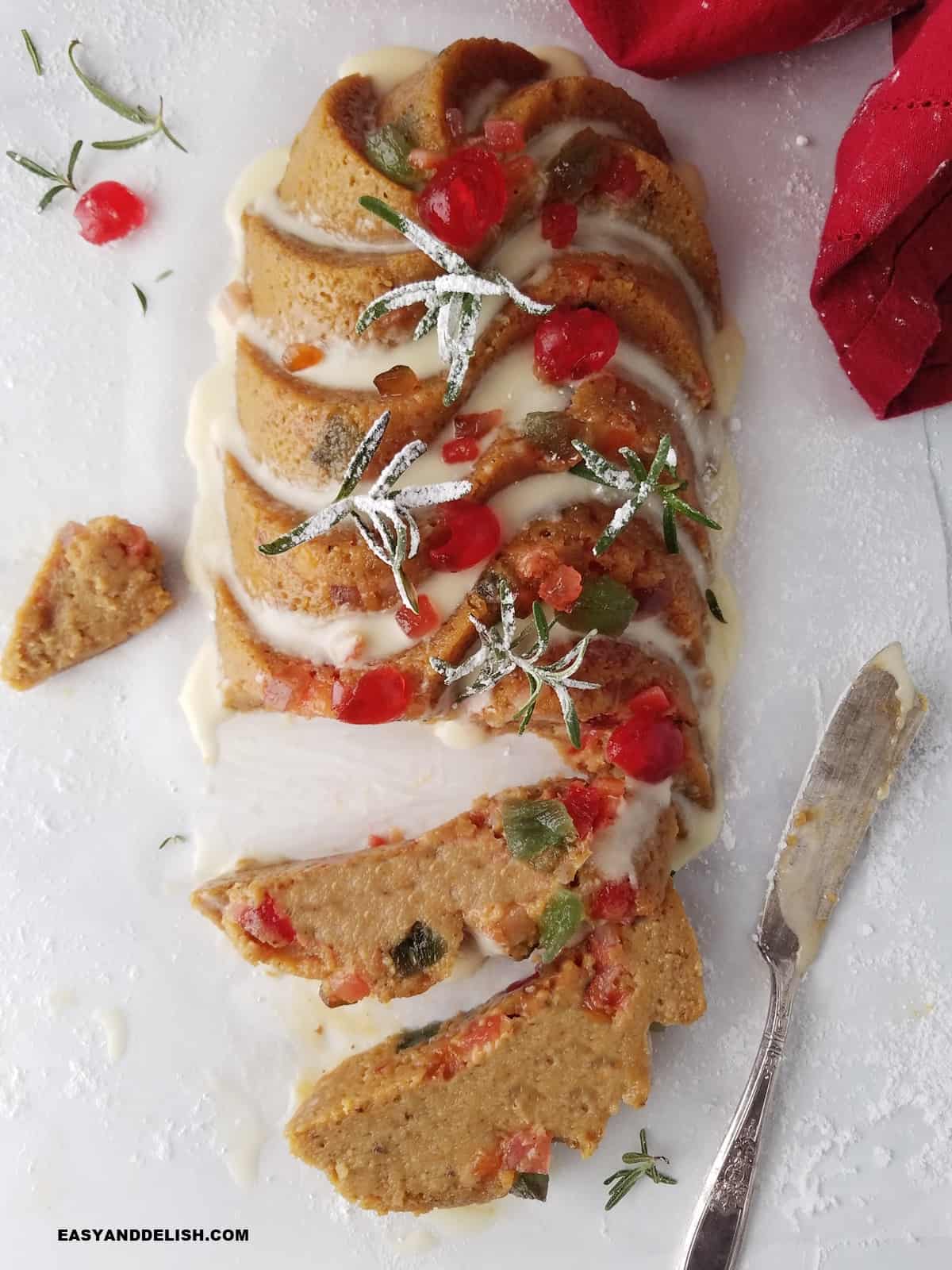 sliced christmas fruitcake with orange icing, decorated with candied fruits and rosemary sprigs.