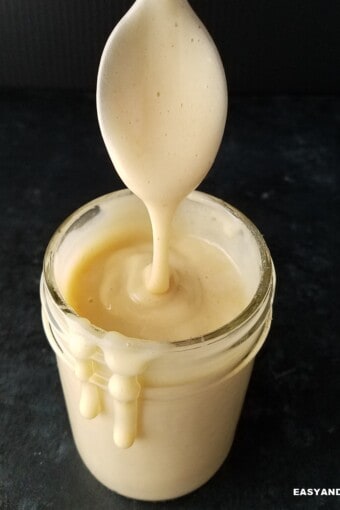 close up of a jar of sugar-free keto condensed milk dripping on the sides with a spoon held on top.