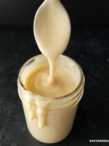 close up of a jar of sugar-free keto condensed milk dripping on the sides with a spoon held on top.